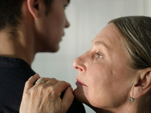 Elderly woman gently holds young man's shoulder, gazing thoughtfully. Soft lighting, neutral background, conveys warmth and connection.