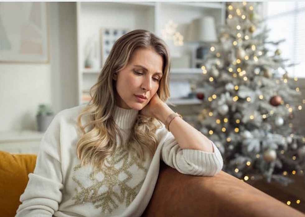 Woman in white sweater looks pensive on a couch. A decorated Christmas tree with white lights is in the background, creating a cozy feel.