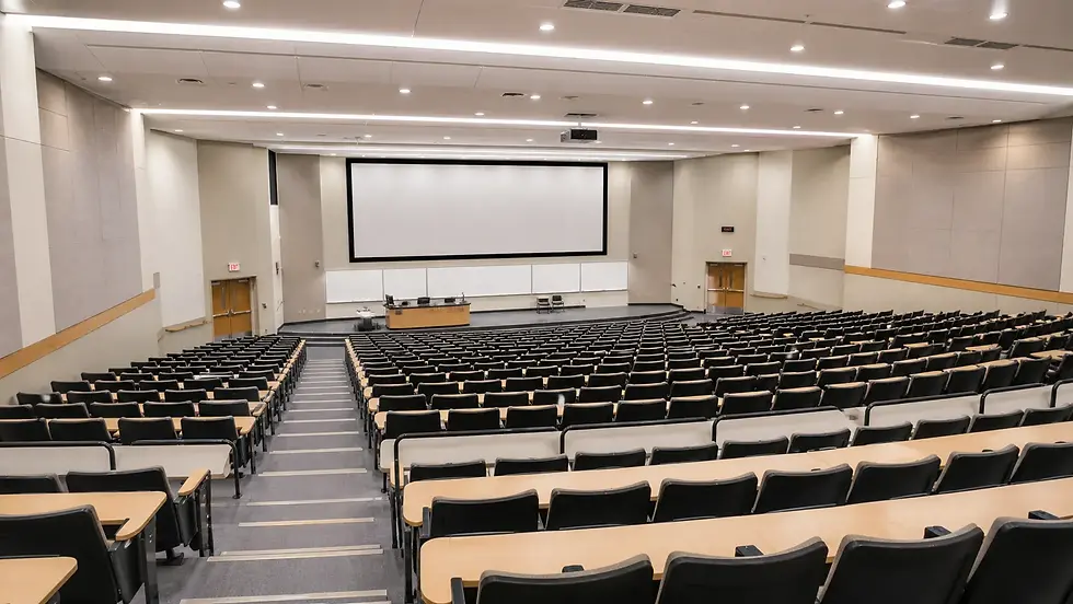 Large empty university lecture hall with tiered seating, rows of desk chairs, a wide projection screen, and a front teaching area.