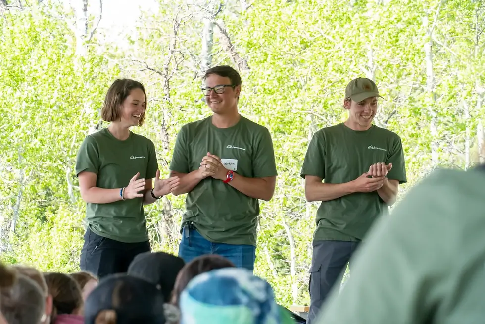 Three OFY leaders in green shirts smiling and clapping while standing on a stage in front of a group of youth outdoors.