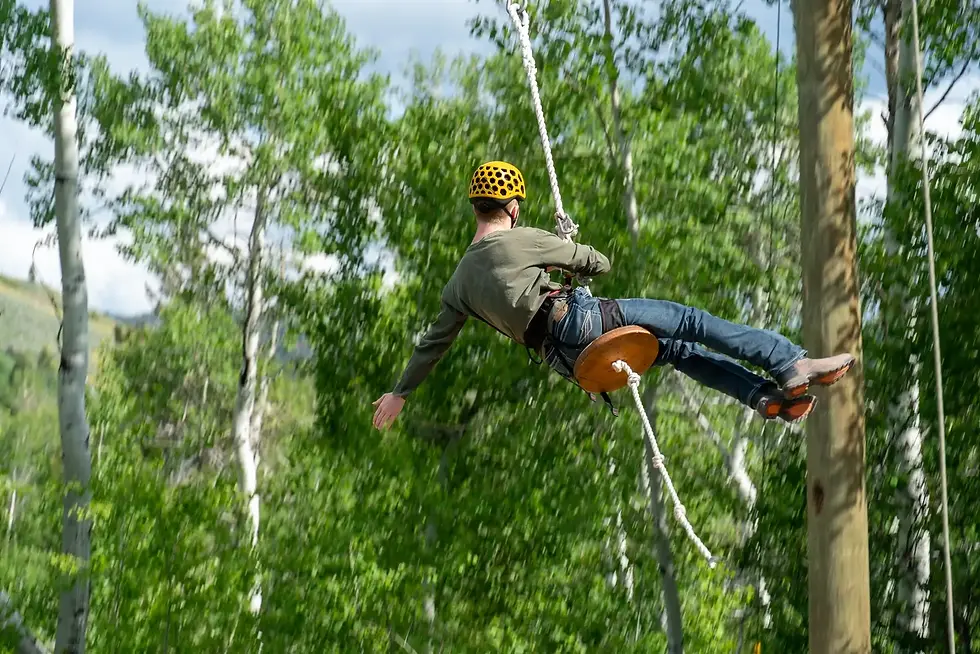Person wearing a helmet swinging from a rope on an outdoor challenge course surrounded by trees.