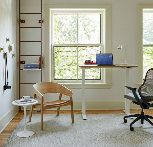 一個辦公或書房空間。窗邊有一張可調節高度的升降桌,搭配一張黑色辦公椅。旁邊則有一張木質的單人椅和一張小邊桌,整體風格實用且自然。