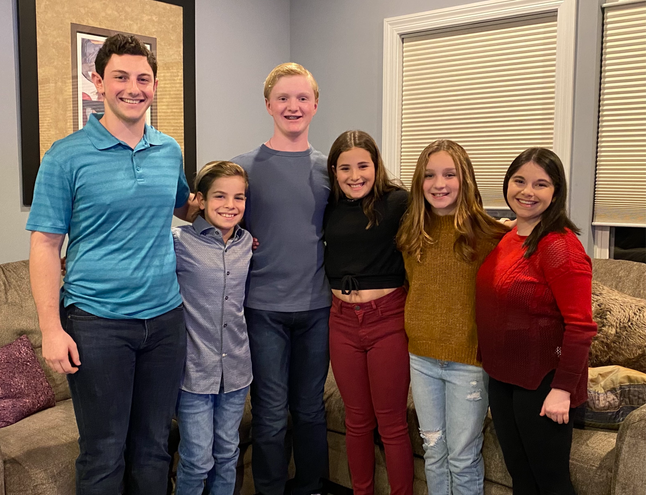 Six kids standing in a line with their arms around each other in front of a light brown living room couch. The three boys on the left are coordinating with shades of blue, while the girls on the right are coordinating with browns, reds, and blacks.