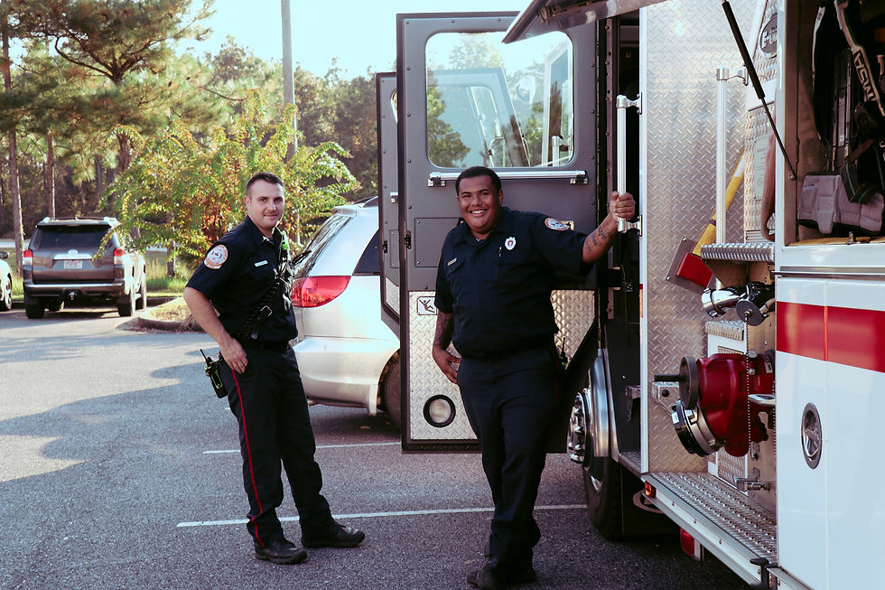 Two firefighters in uniform stand by an open fire truck in a sunny parking lot, smiling and relaxed. Trees and parked cars in the background.