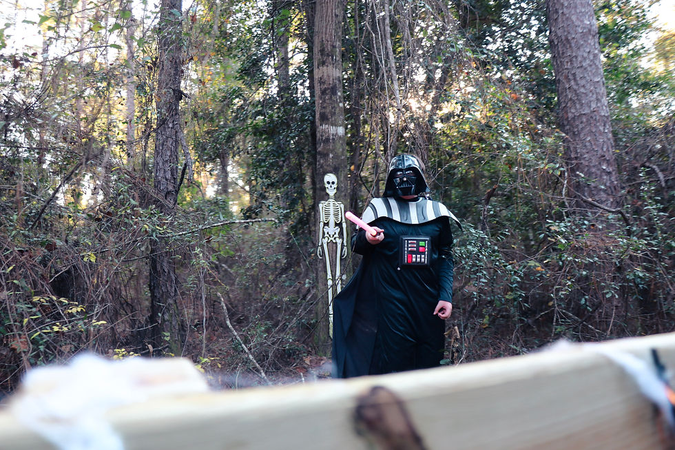 Person in a Darth Vader costume with a pink lightsaber stands in a forest beside a skeleton decoration, wearing a black cape.