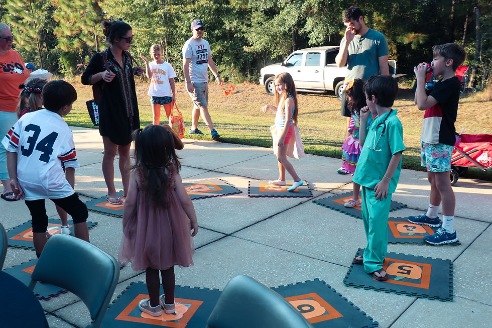 Kids play on pumpkin mats at an outdoor party. Adults supervise. It's sunny and trees line the background, creating a joyful atmosphere.