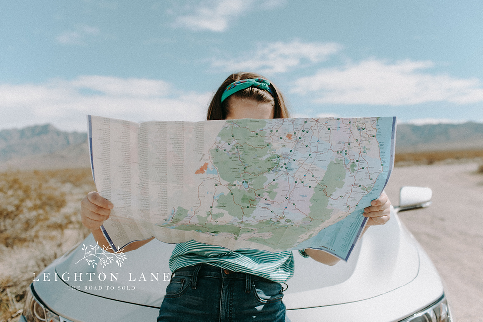 Girl studies a folded road map beside a white car on a desert highway under blue sky; Leighton Lane logo visible.