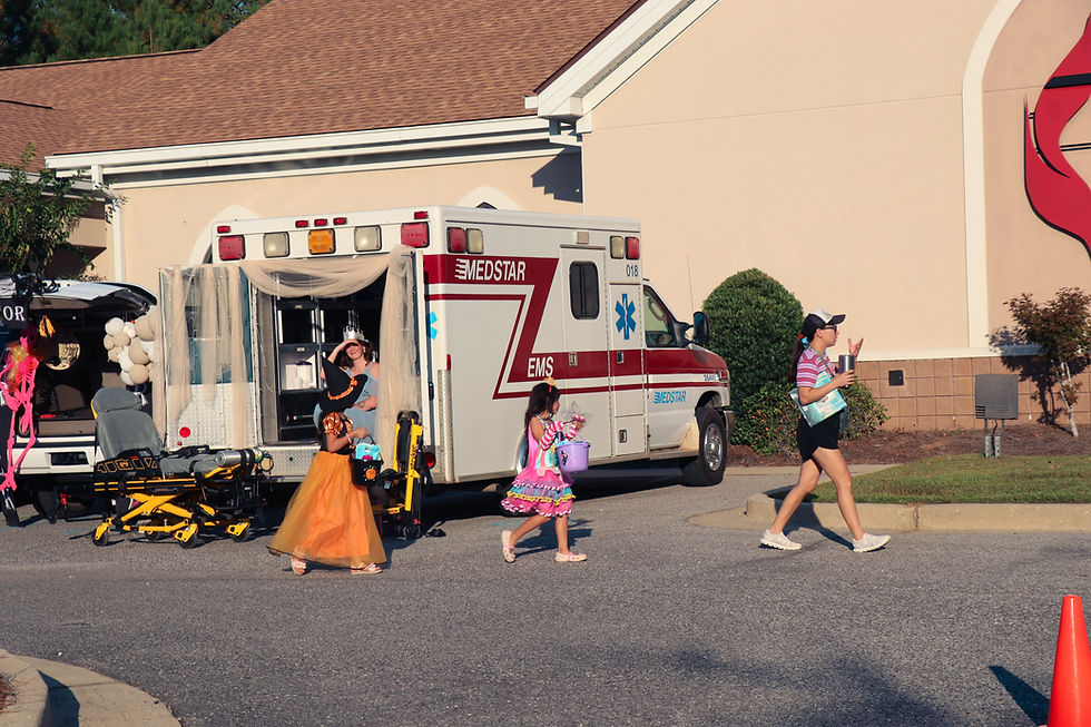 Kids in colorful costumes walk past an ambulance with open doors and medical staff inside. A woman waves, carrying a cup, under a sunny sky.