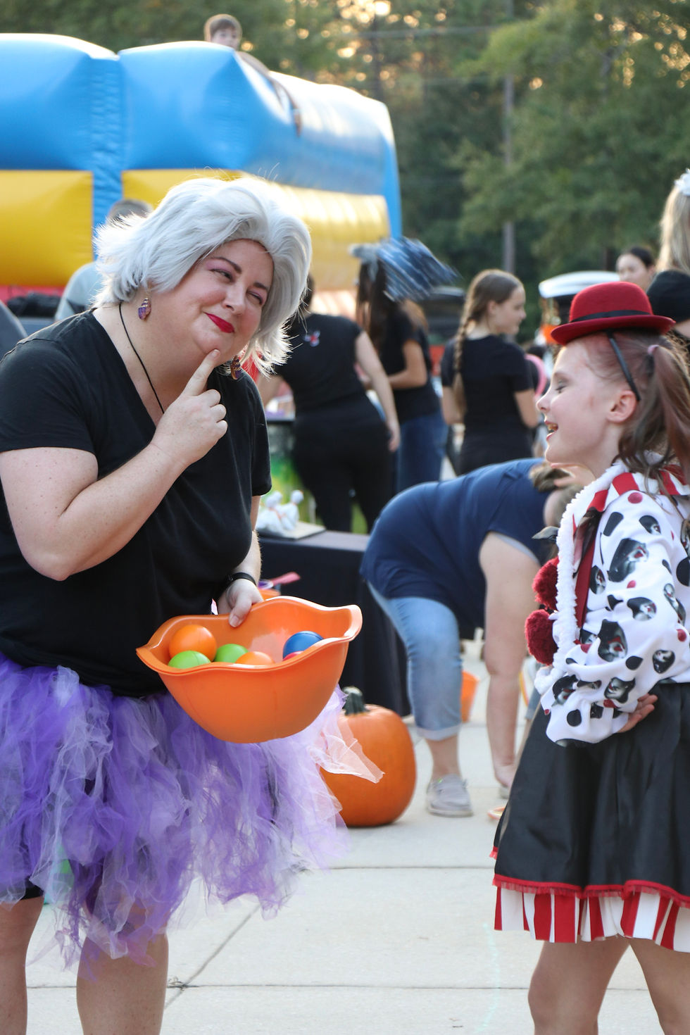 Person in a purple tutu holds an orange bowl of balls, smiling at a child in costume. Outdoor event with colorful inflatable in background.