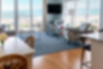 Bright living room with beach view, floral armchairs, a blue rug, and white furniture. Ocean in the background through large windows.