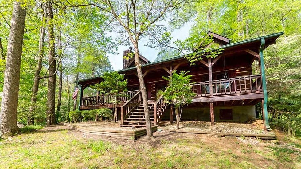 Wooden cabin with a porch set in a lush green forest. Steps lead up to the entrance. Sunlight filters through the leafy trees.