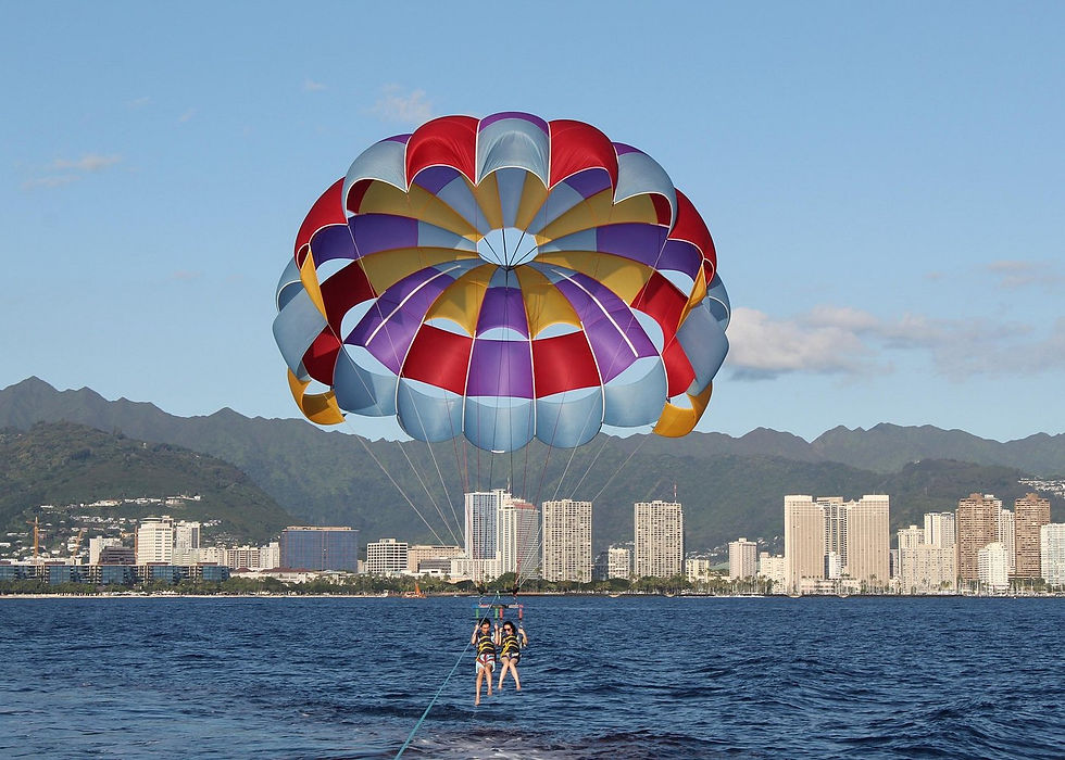 Two parasailers glide over the ocean beneath a colorful canopy, with a city skyline and mountains behind.