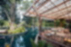 Woman in white dress sits at a wooden table on a patio by a reflective pool, surrounded by lush greenery and trees, under a rustic awning.