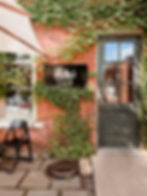 Brick wall with ivy, a door, a window, and a "Rubi's Cafe" sign. Black chair on stone patio, creating a cozy, rustic vibe.