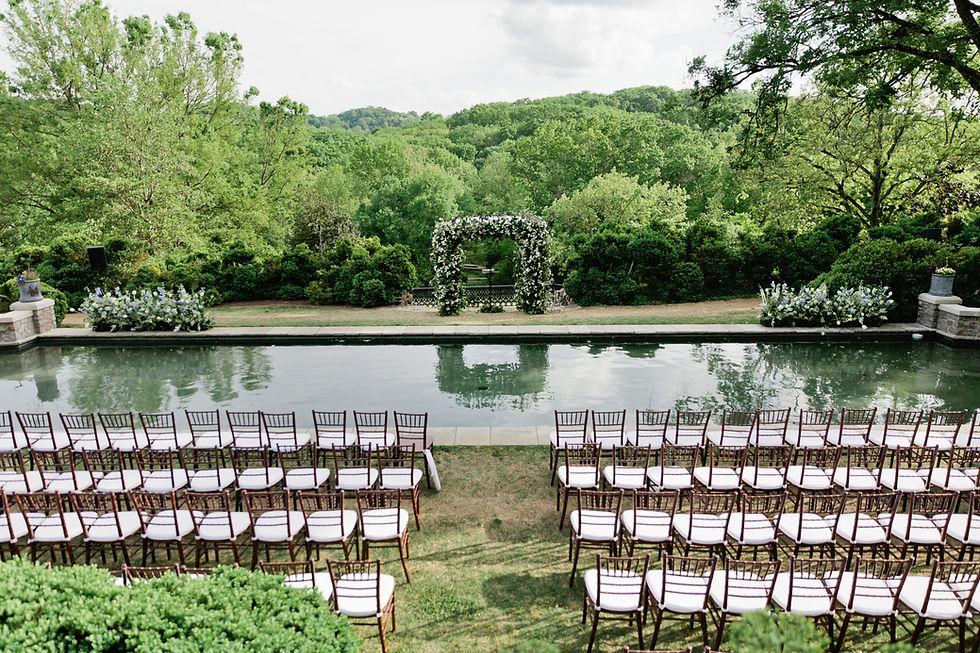 Wedding setup with rows of brown chairs by a reflective pond. A floral arch is centered against a lush green forest backdrop. Peaceful mood.
