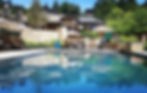 A serene poolside scene with lounge chairs, umbrellas, and a stone building. Green trees surround the area, reflecting in the water.