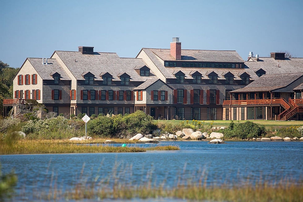 Large shingled lodge with red shutters beside a calm lake, framed by rocks and grasses under a clear blue sky.