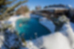 People swim in a heated pool surrounded by snow and icicles. Clear blue sky and glass-walled building in the background on a sunny day.