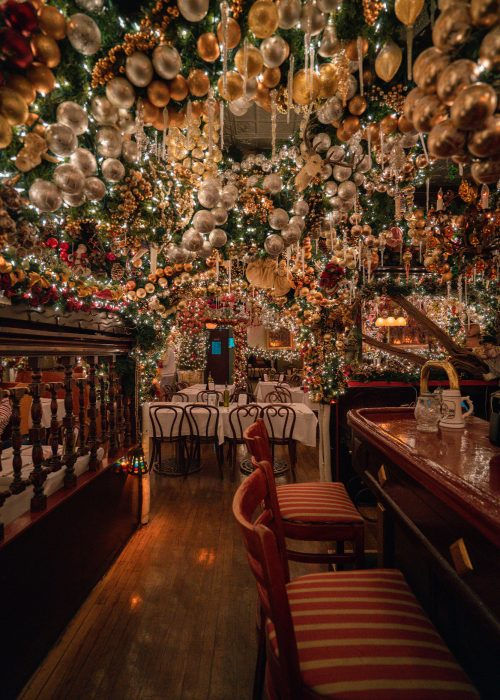 Festive restaurant with lush Christmas decorations, gold and silver ornaments, twinkling lights, empty tables, wooden bar, and red-striped chairs.