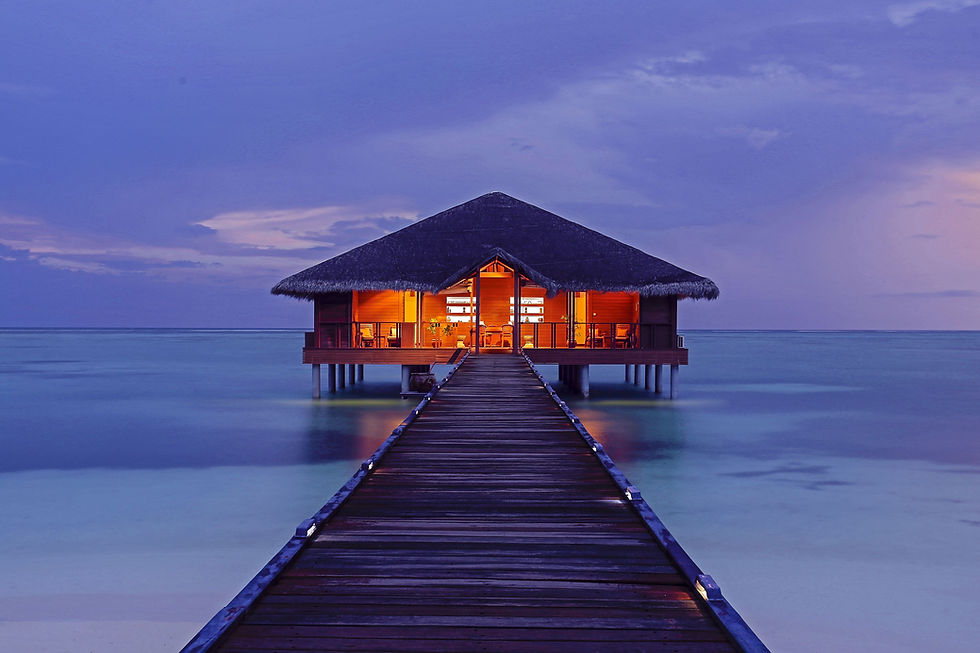 Tropical hut over water at dusk, illuminated warmly, with a wooden walkway. Calm sea surrounds, under a vibrant purple-blue sky. Peaceful mood.