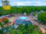 Aerial view of an amusement park on a cloudy day. Yellow gondola, people walking near a central fountain, surrounded by trees and colorful shops.