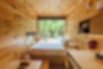 Cozy wooden cabin interior with a bed by a large window. Views of green forest outside; red phone, shelves, and soft lighting inside.