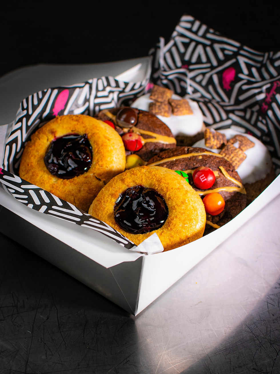 A box with colorful cookies on black-and-white zigzag paper. Two cookies have dark jam, surrounded by a dark surface. Bright, inviting mood.