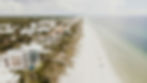 Aerial view of beachfront homes and people enjoying a sunny day on a sandy beach. Turquoise ocean on the right and greenery in the background.