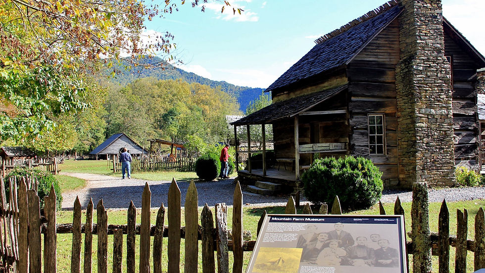 Historic log cabin at Mountain Farm Museum with two people and lush trees. Wooden fence in foreground, sunlit and scenic.