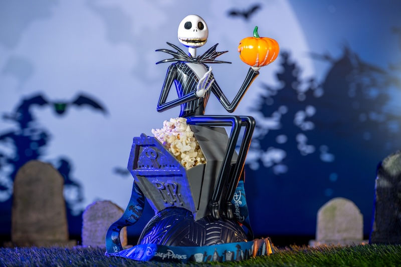 Skeleton figure holds a pumpkin and popcorn against a blue, moonlit graveyard backdrop with bats. The setting is whimsical and eerie.