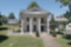 White colonial house with large columns, porch, and American flag. Green lawn and trees surround, under a clear blue sky. Peaceful setting.