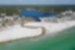 Aerial view of a beach with white sand, emerald water, and houses lining a bay. People relax on the shore under a partly cloudy sky.