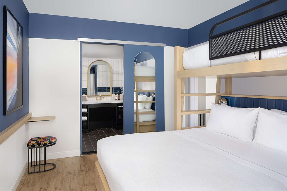 Modern hotel room with a bunk bed, blue and white decor, and wood flooring. A colorful stool and bathroom with a gold mirror in view.