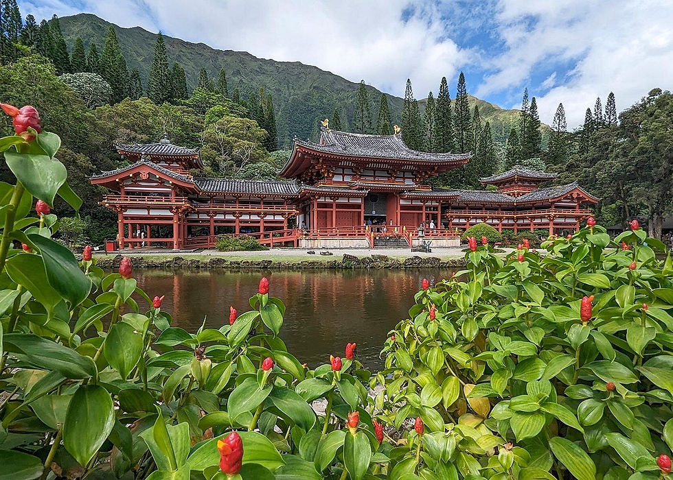 Red temple beside a pond, framed by green flowers, with tall pines and mountains under a cloudy blue sky.