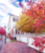 White house with black shutters, surrounded by vibrant autumn trees and a white fence. Red brick path, under a bright blue sky.