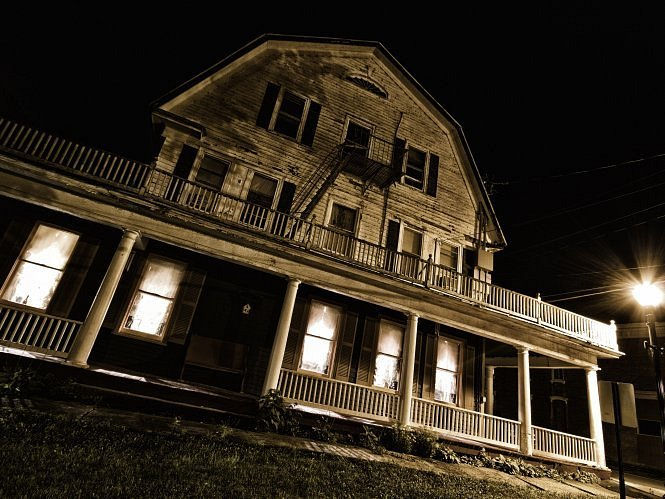 A dimly lit, old wooden house at night, with glowing windows and a streetlamp nearby, creates a mysterious and eerie atmosphere.