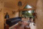 Cozy wooden cabin interior with a gray sofa, tennis racquets on the wall, and a view of a deck and trees. Warm lighting and a vintage rug.