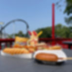 A table with hot dogs, a corn dog, Fritos, and a colorful milkshake. Roller coaster and trees in the background under a clear blue sky.