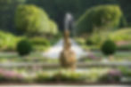 Garden with central stone fountain spraying water, surrounded by colorful flowers and neatly trimmed hedges, with tree-lined path behind.