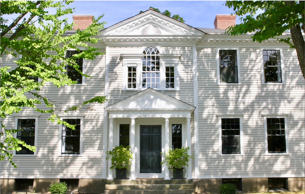 White colonial-style house with a black door, flanked by trees on a sunny day. Shadows and greenery add contrast to the serene setting.