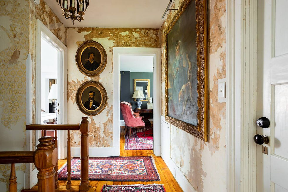 Hallway with peeling wallpaper, two oval portraits, and a large painting. Red rugs cover wooden floors. A room with a red chair is visible.