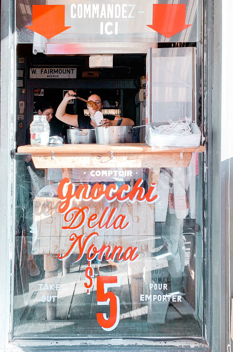 Woman serving pasta in a shop. Large red and white text on glass reads "Gnocchi Della Nonna $5." Bright, casual setting.