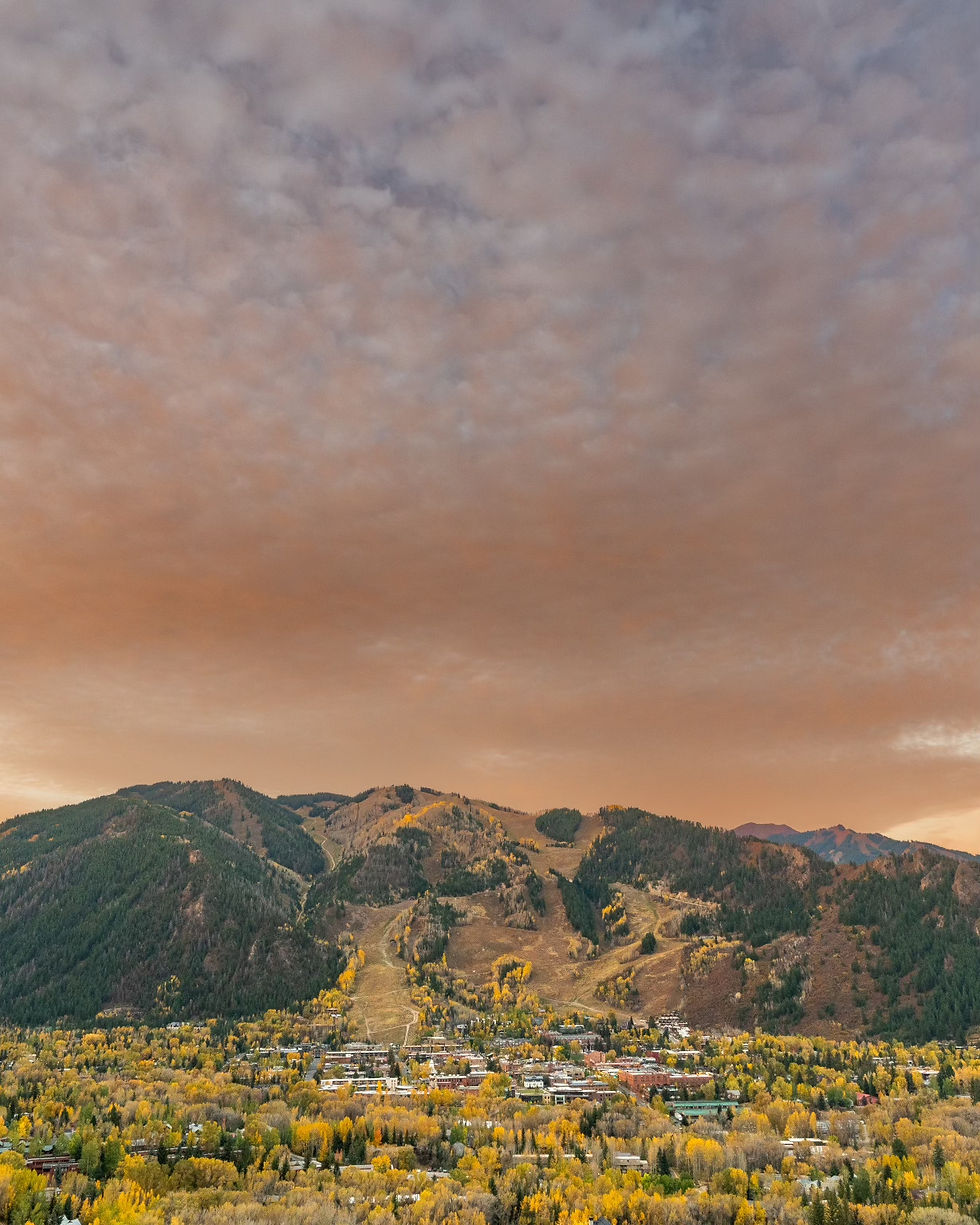 A serene autumn landscape of a town with colorful trees, nestled below green and brown mountains under a cloudy, orange-tinted sky.