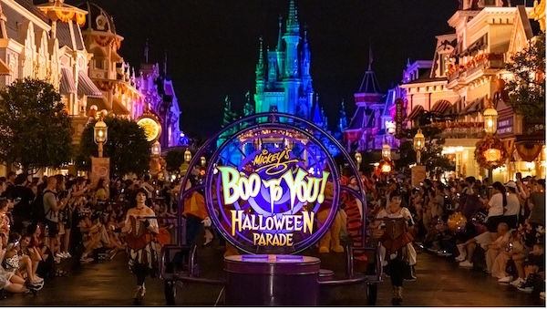 Halloween parade with dancers in festive costumes at night. Bright sign reading "Mickey's Boo to You Halloween Parade" amid colorful lights.