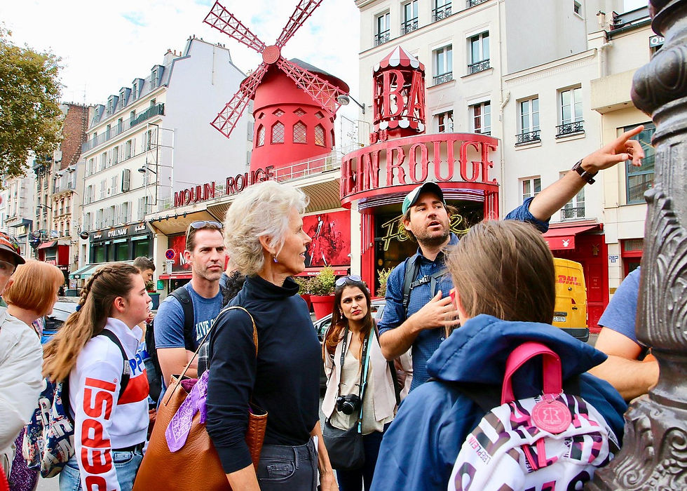 Tourists listen to a guide outside the red Moulin Rouge windmill in Paris, with the sign and street buildings visible.