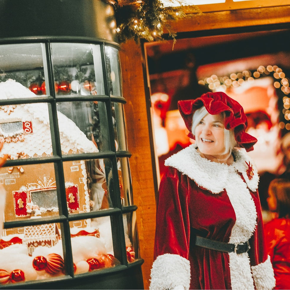 Woman in red Santa outfit smiles beside a window showcasing a gingerbread house. Warm lights and festive decor in the background.