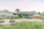 Beachfront resort with wicker loungers, grassy foreground, and elegant white buildings in the background. Clear sky, serene ambiance.