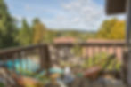 Balcony view with two colorful chairs, a small table holding a lamp and plant. Overlooks trees, rooftops, and a pool under clear skies.