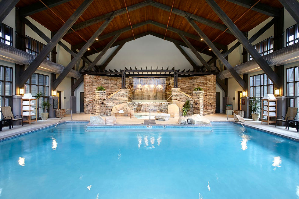 Indoor pool with blue water, surrounded by stone walls and wooden beams. A waterfall feature is in the center. Calm, inviting atmosphere.