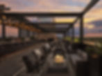 Rooftop bar at sunset with string lights, modern seating, and plants. Warm colors in the sky create a cozy, inviting atmosphere.